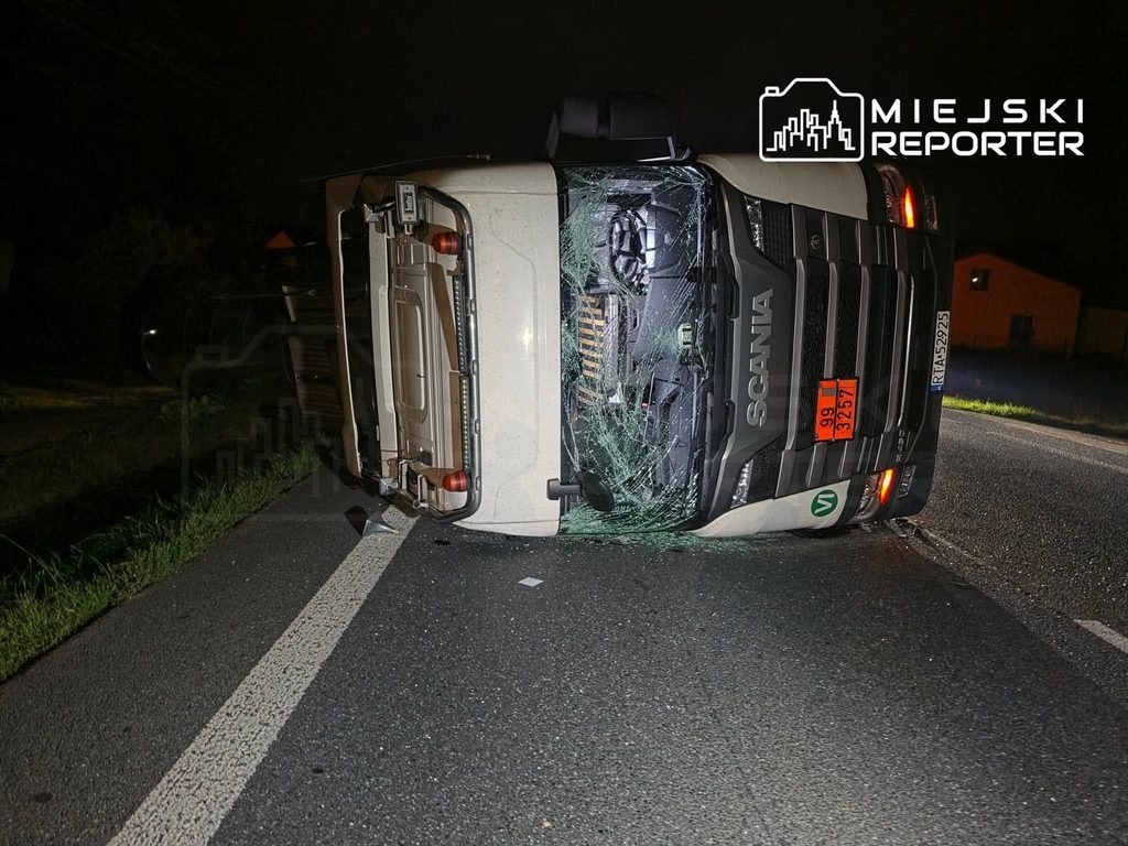 Cysterna marki Scania przewrócona na drodze, z uszkodzoną kabiną i rozbitą szybą, w ciemnej scenerii nocnej.