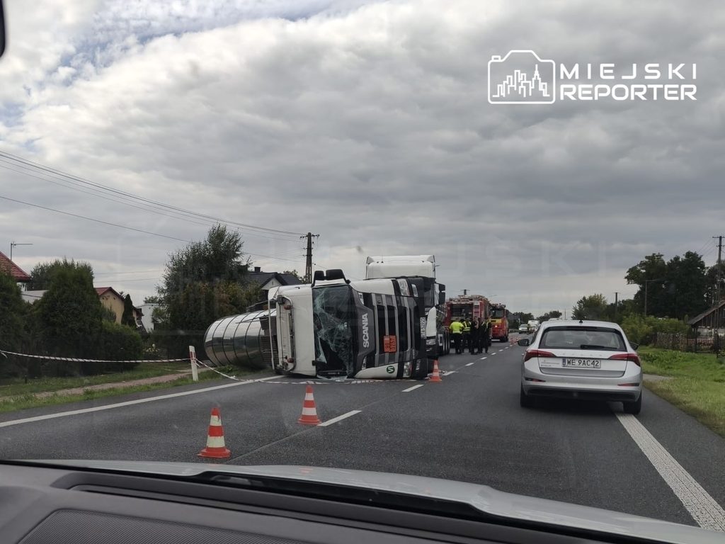 Cysterna z asfaltem przewrócona na drodze, obok stoją pojazdy służb ratunkowych, a w tle widać zabudowania. Utrudnienia na DK50.