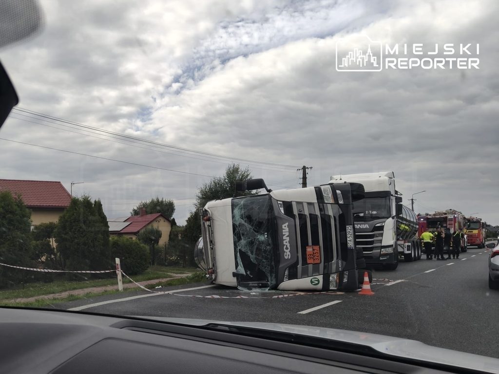 Cysterna z asfaltem przewrócona na jezdni, obok pracują strażacy i służby drogowe, w tle domy i chmurne niebo. Utrudnienia na DK50.