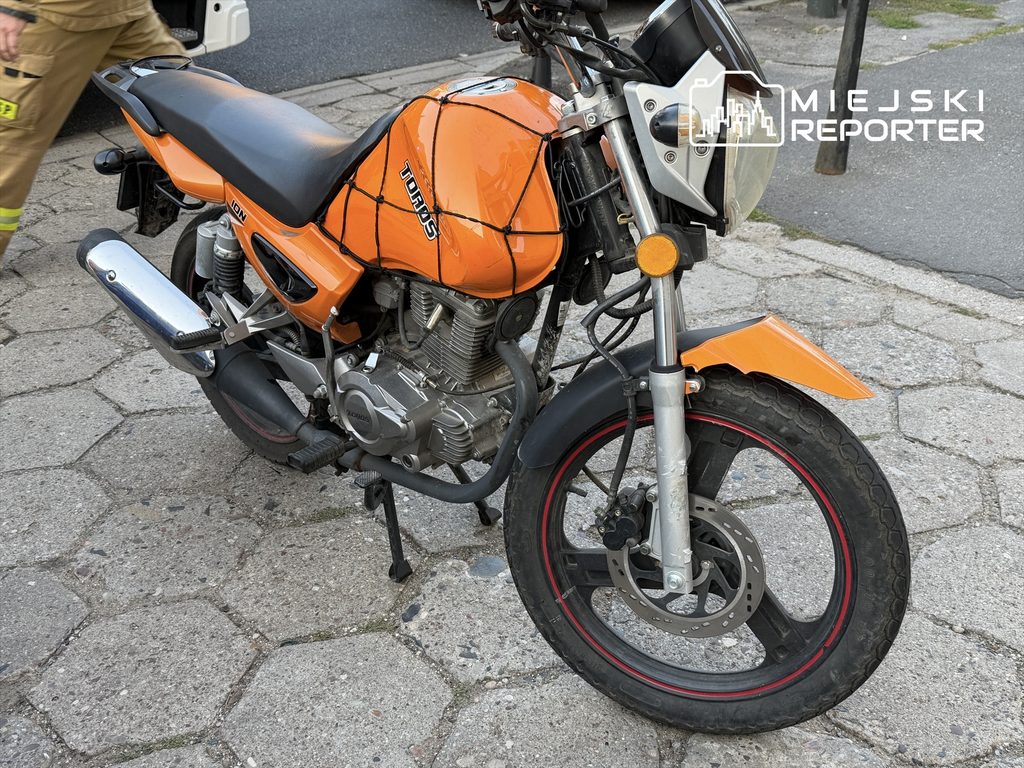 Motocykl w pomarańczowym kolorze zaparkowany na chodniku, obok widoczne płyty chodnikowe.