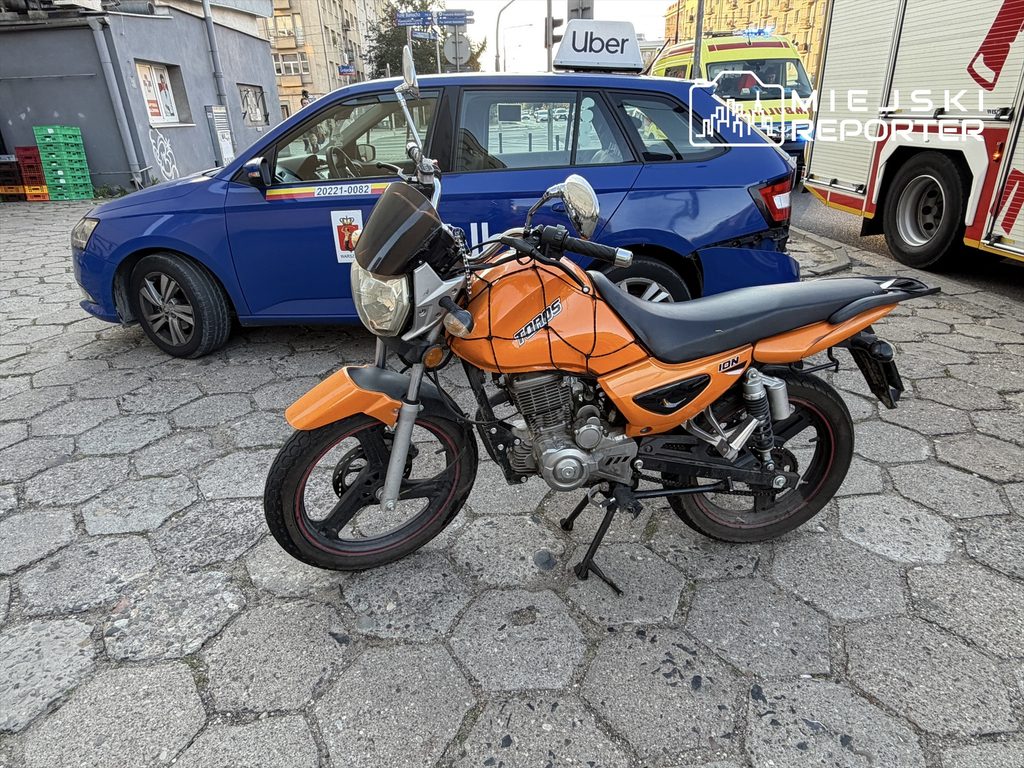 Motocykl w pomarańczowym kolorze stoi na kostce brukowej obok niebieskiego samochodu z napisem „Uber”.