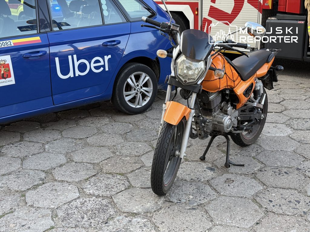 Motocykl w pomarańczowym kolorze stoi na chodniku obok niebieskiego samochodu z logo Ubera.