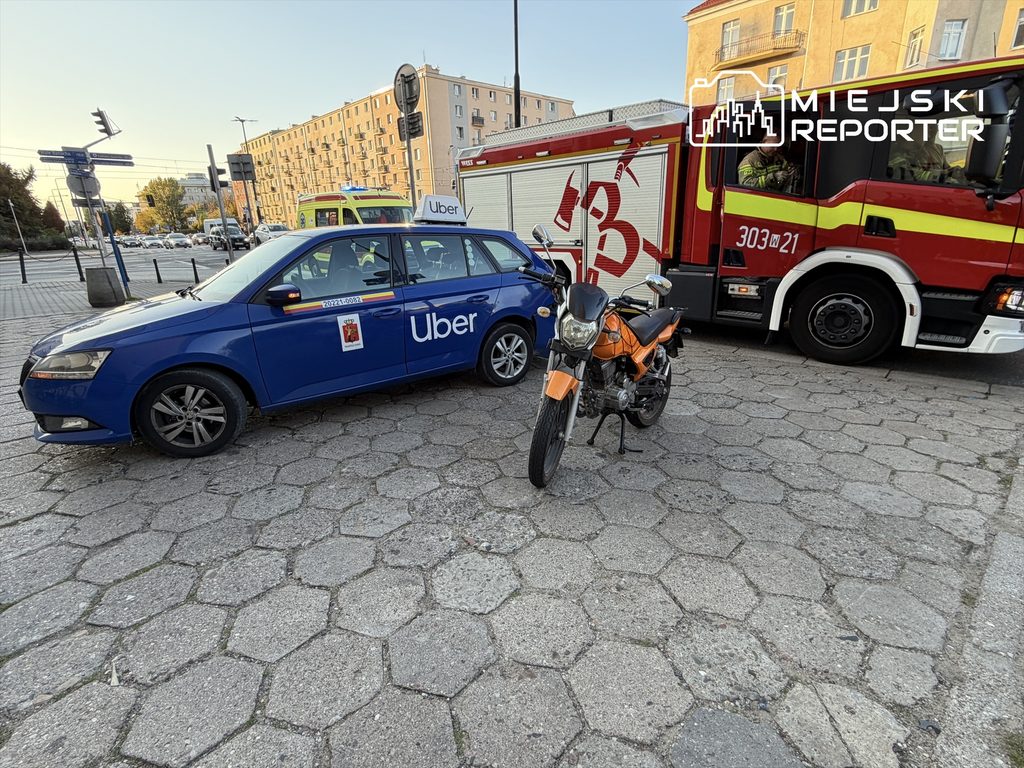 Nieoznakowany samochód osobowy marki Skoda z napisem "Uber" stoi obok pomarańczowego motocykla, a w tle widoczny jest wóz strażacki.