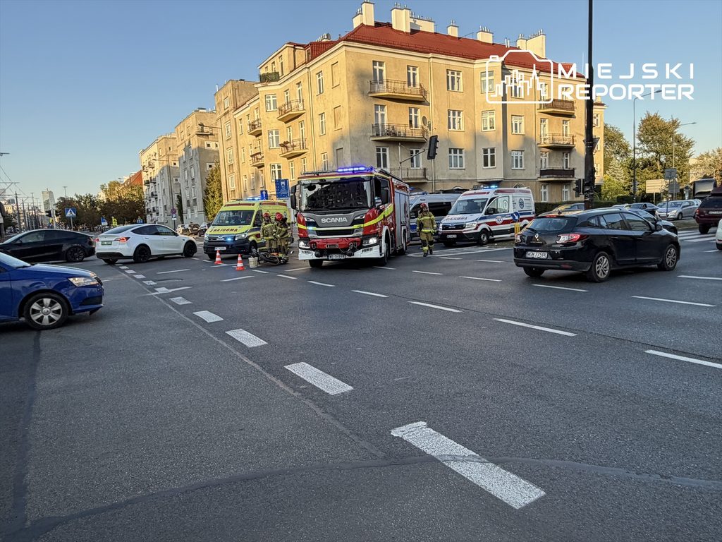 Na skrzyżowaniu ulicy stoją pojazdy służb ratunkowych, w tym straż pożarna i karetka, obok zderzone auto i motocykl.