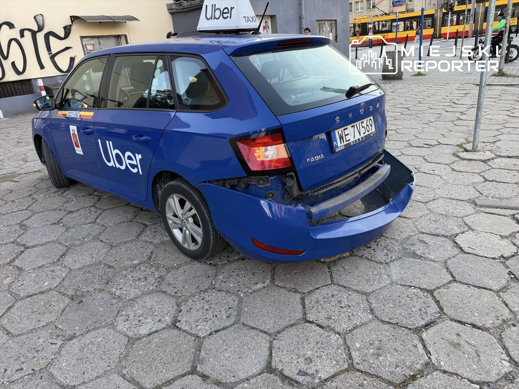 Uszkodzony niebieski samochód marki Skoda z widocznym uszkodzeniem tyłu, zaparkowany na chodniku.