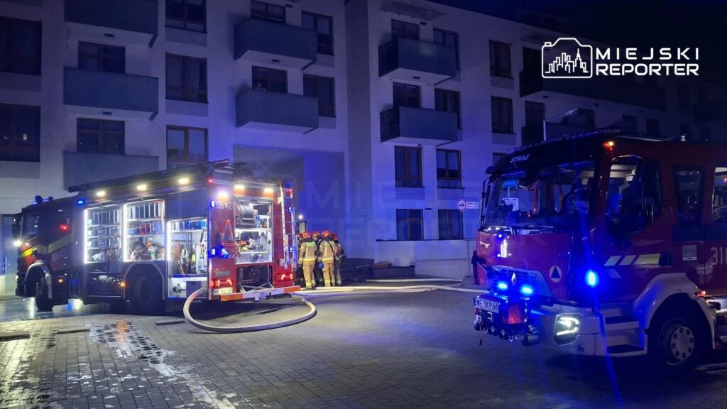 Strażacy w mundurach przygotowują sprzęt przy wozie strażackim przed nowoczesnym budynkiem mieszkalnym.