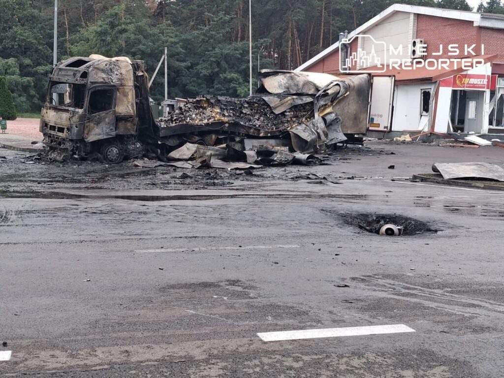 Spalony tir leży na zniszczonym placu, w tle widoczny uszkodzony budynek.