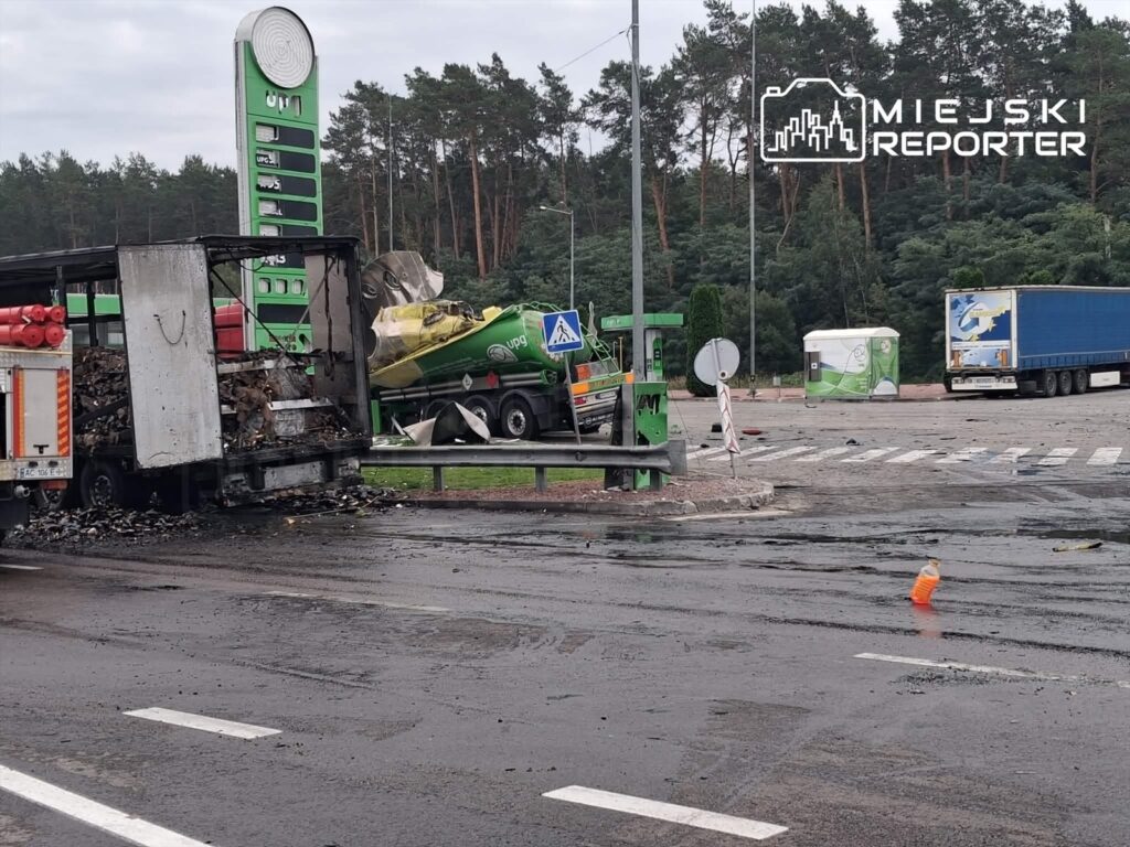 Zniszczony samochód ciężarowy z palącym się ładunkiem stoi na drodze obok stacji paliw w lesie.