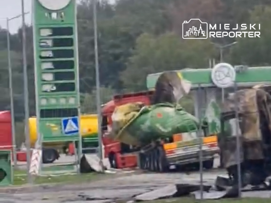 Zniszczony parking z przewróconymi ciężarówkami i uszkodzoną stacją paliw w tle.