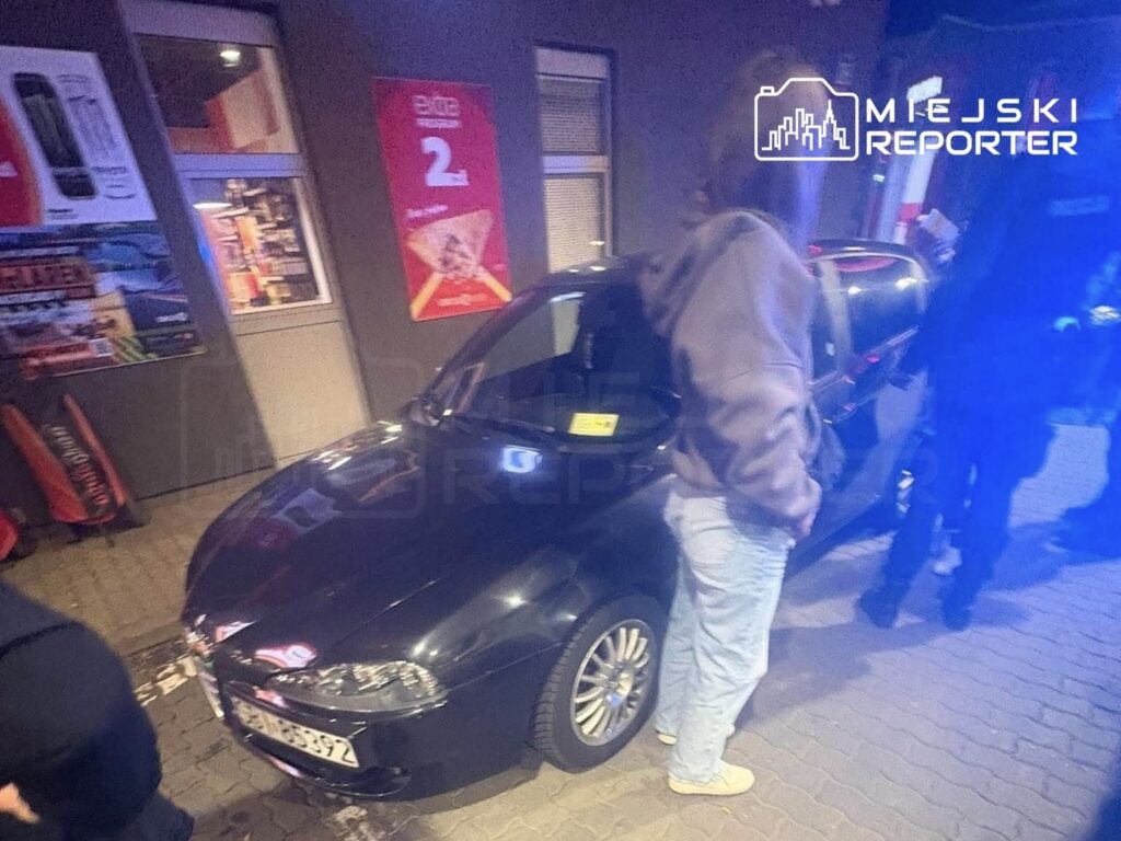 Mężczyzna w szarej bluzie stoi obok czarnego samochodu zaparkowanego przed budynkiem z reklamami.