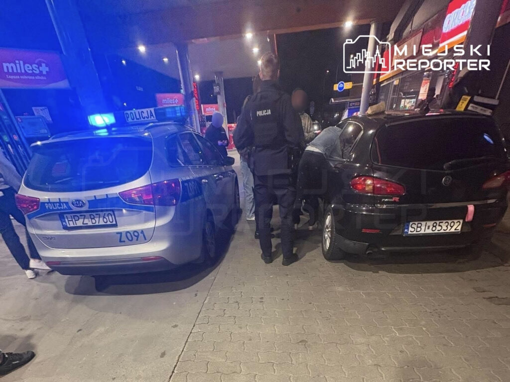 Policjanci w mundurach interweniują przy stacji benzynowej, obok dwóch zaparkowanych samochodów.
