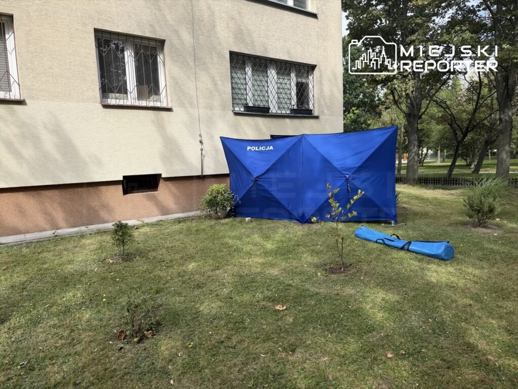 Namiot policyjny rozstawiony w zielonym terenie przed blokiem mieszkalnym, obok leży niebieska torba.