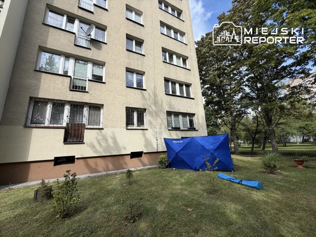 Mundurowi zabezpieczają miejsce zdarzenia przy bloku, obok niebieskiego namiotu policyjnego na trawie.