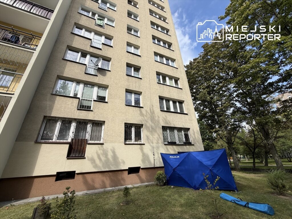 Mężczyzna leży pod niebieskim namiotem policyjnym przy bloku mieszkalnym, otoczony przez zieloną trawę i drzewa.