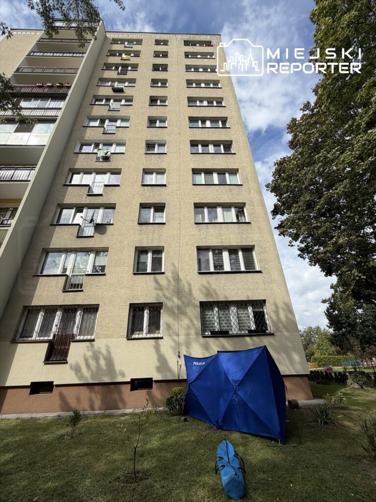 Mężczyzna w niebieskim namiocie policyjnym stoi obok bloku mieszkalnego w otoczeniu zieleni.