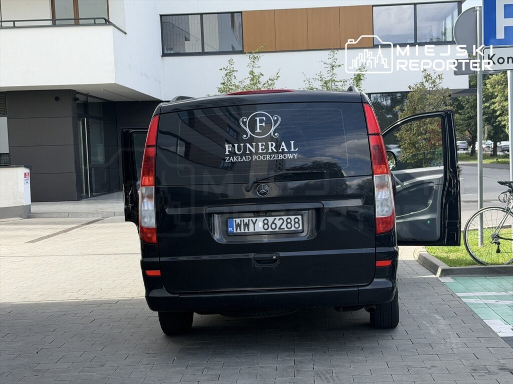 Czarny samochód z logo "FUNERAL ZAKŁAD POGRZEBOWY" zaparkowany przed nowoczesnym budynkiem, z otwartymi drzwiami tylnymi.