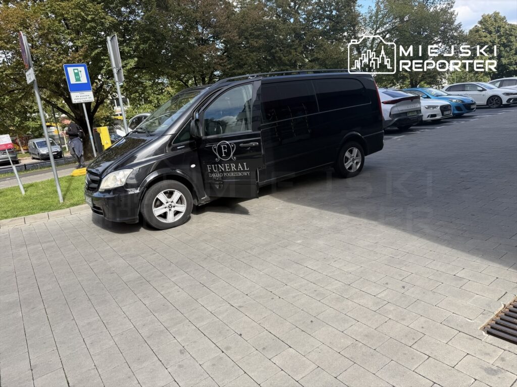 Czarny samochód pogrzebowy zaparkowany na chodniku, obok znaków drogowych i zielonej trawy.