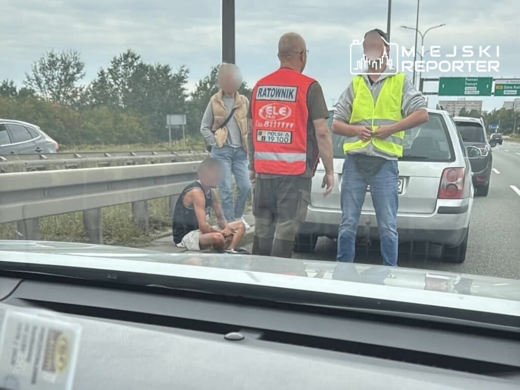 Mężczyzna w krótkich spodniach siedzący na poboczu drogi, obok dwóch osób w odblaskowych kamizelkach, samochody w tle.