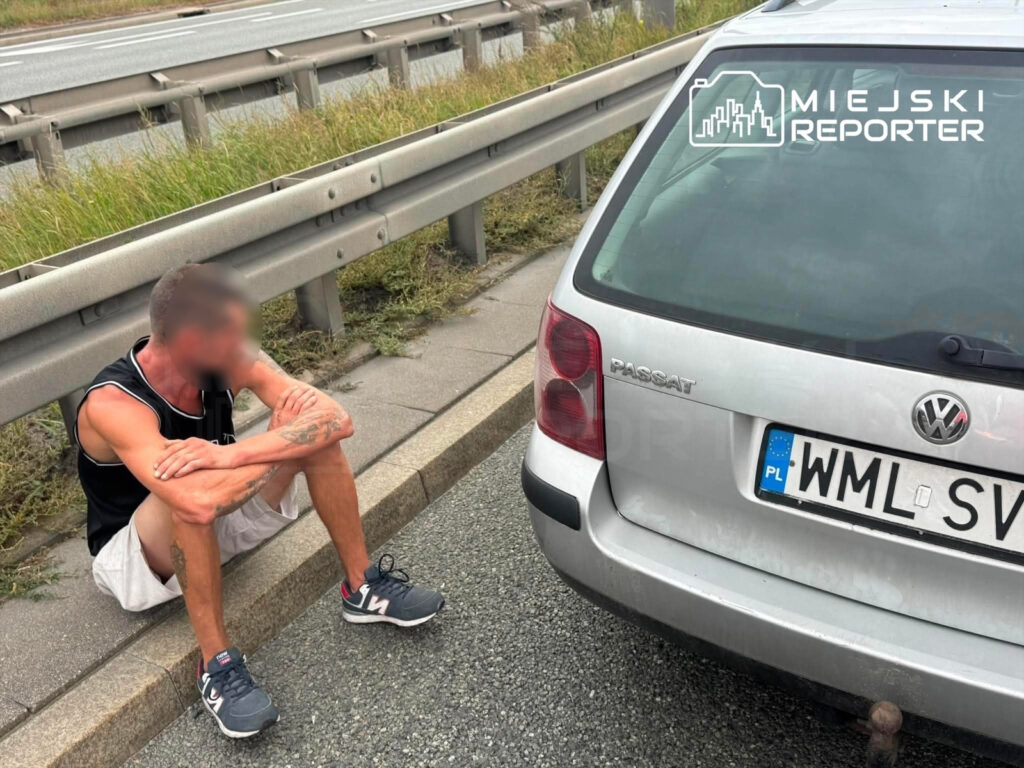 Mężczyzna w sportowym stroju siedzący na poboczu drogi obok zaparkowanego samochodu osobowego.
