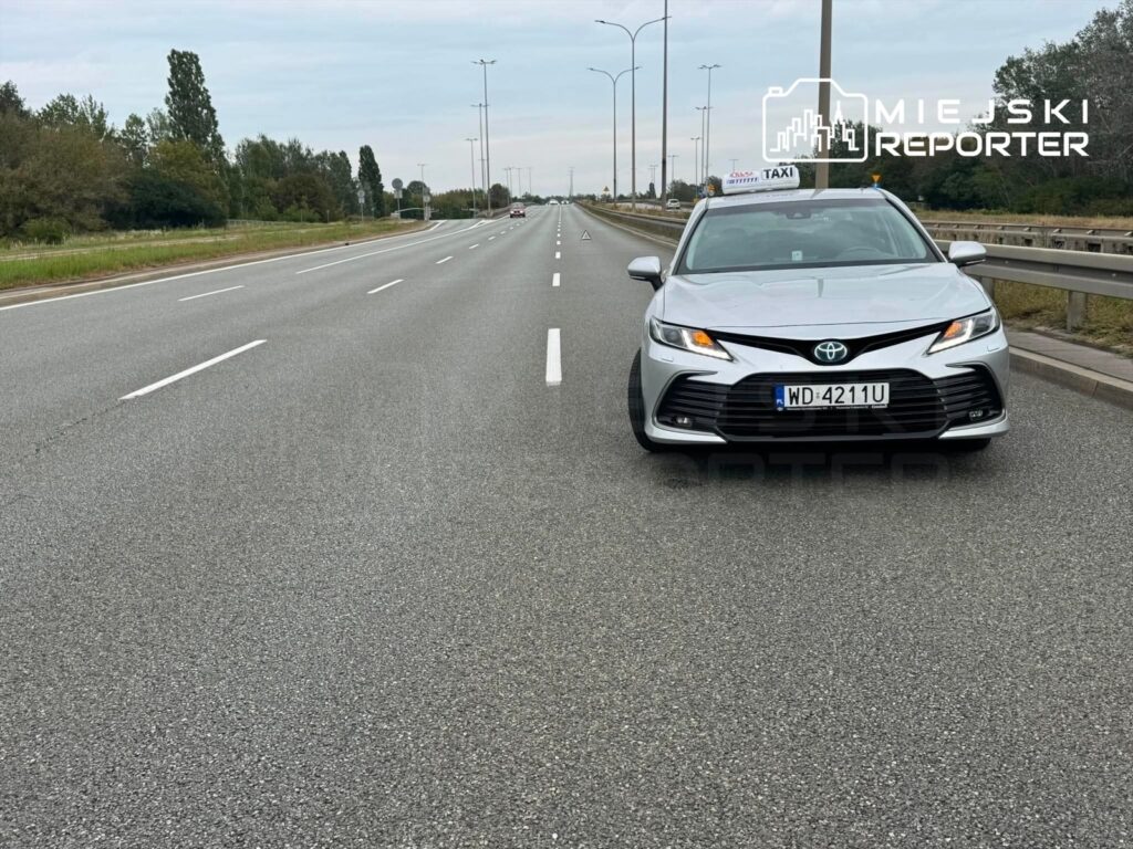 Taksi stojące na pustej drodze, z widocznymi pasami ruchu i drzewami w tle. Na dachu widoczny znak "TAXI".