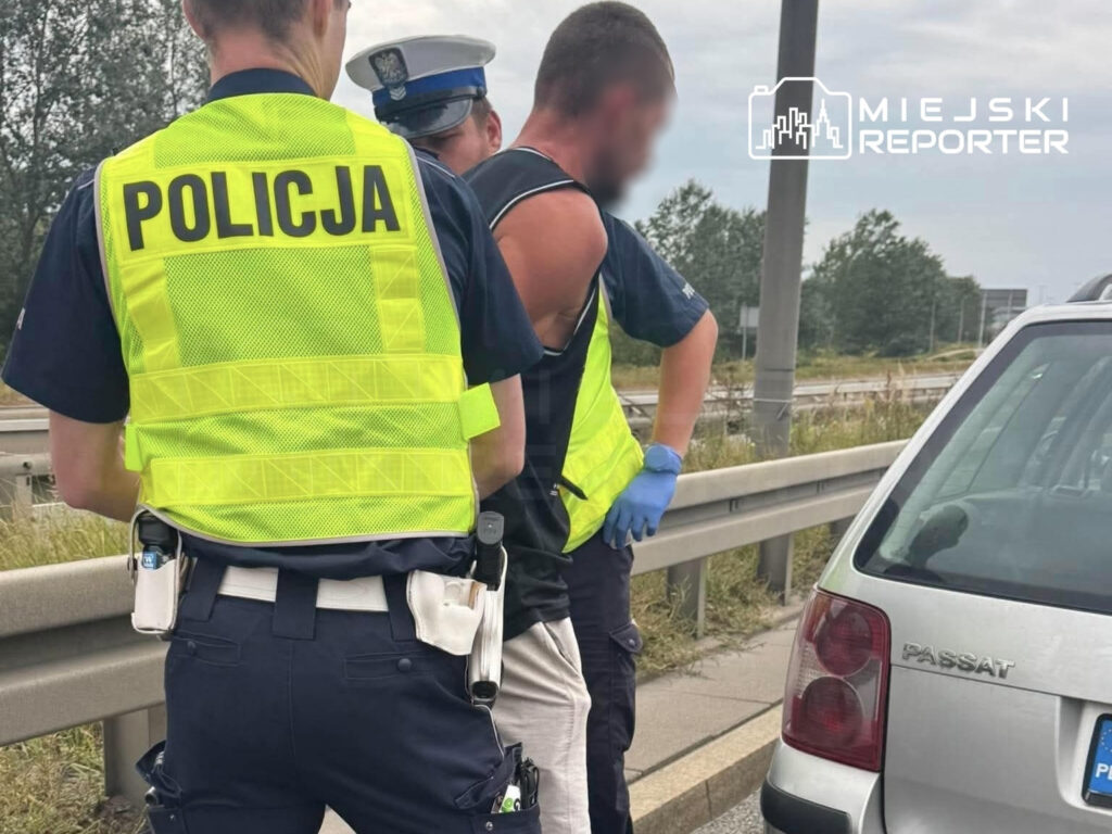 Policjanci w odblaskowych kamizelkach zatrzymują mężczyznę przy Trasie Siekierkowskiej, obok samochodu osobowego.