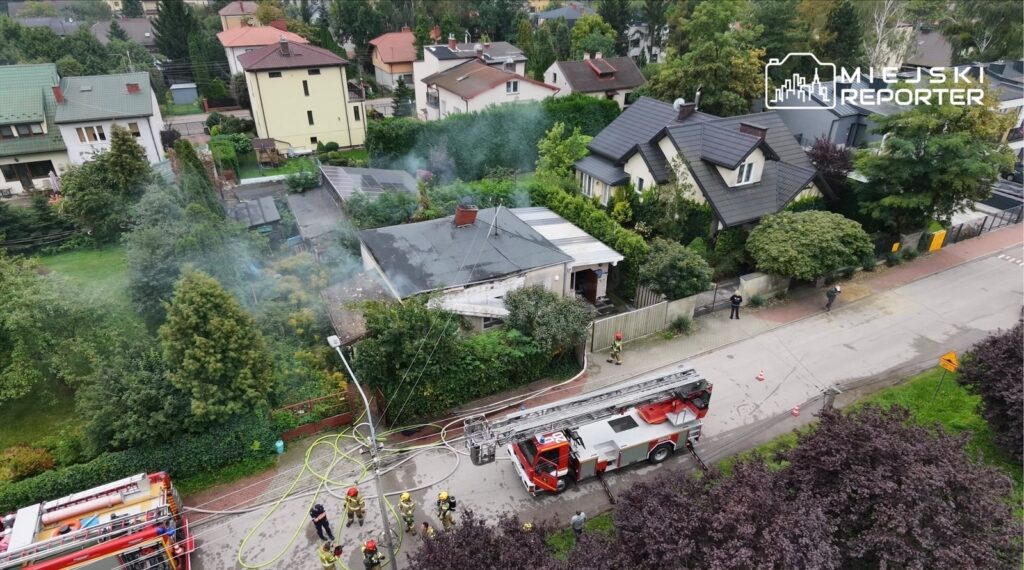 Strażacy z wężami gaśniczymi pracują przy zadymionym budynku mieszkalnym w otoczeniu domów jednorodzinnych.