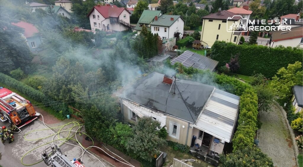 Dym wydobywający się z dachu domu jednorodzinnego w otoczeniu zieleni, obok wóz strażacki i węże gaśnicze.