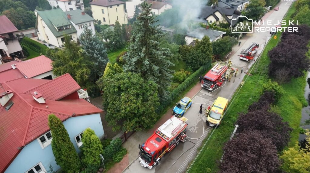 W okolicy domów z dachami pokrytymi czerwoną dachówką, strażacy gaszą pożar, a obok stoi karetka i radiowóz.