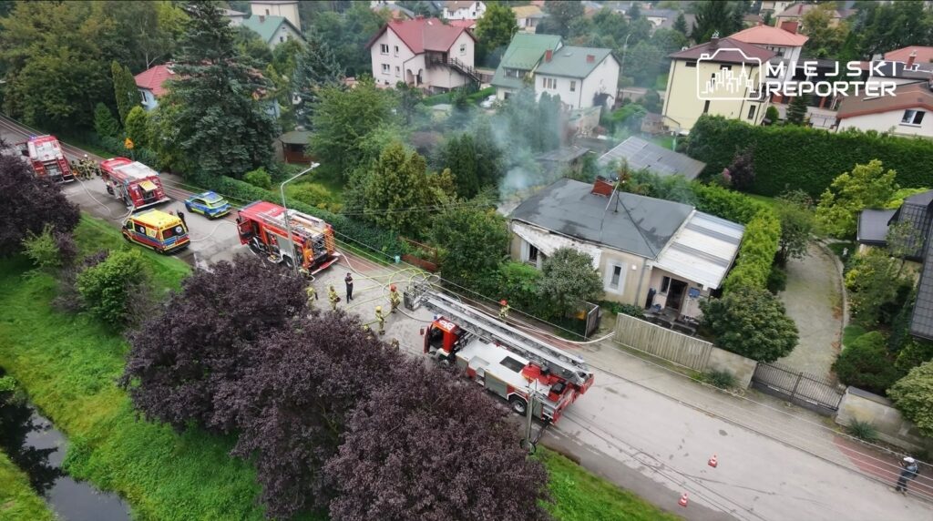 Strażacy gaszą pożar w domu jednorodzinnym, obok zaparkowane wozy strażackie i radiowóz, w tle gęsta roślinność.