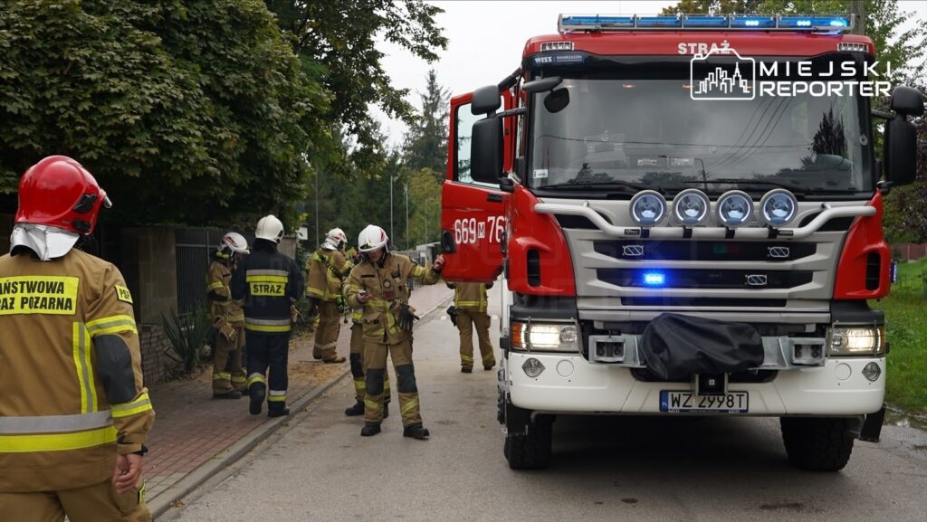 Strażacy w mundurach i hełmach pracują przy wozie strażackim na ulicy, przygotowując się do akcji gaśniczej.