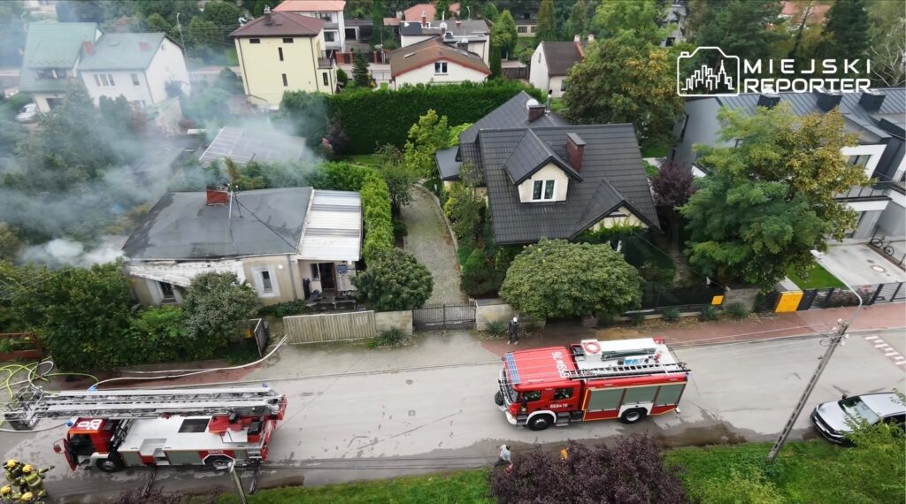 W tle widać dym wydobywający się z dachu budynku, obok stoi wóz strażacki z rozwiniętą drabiną.
