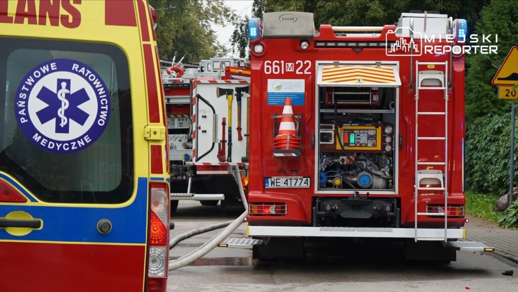 Zespół ratunkowy w karetce z logo "Państwowe Ratownictwo Medyczne" stoi obok wozu strażackiego z otwartą klapą.
