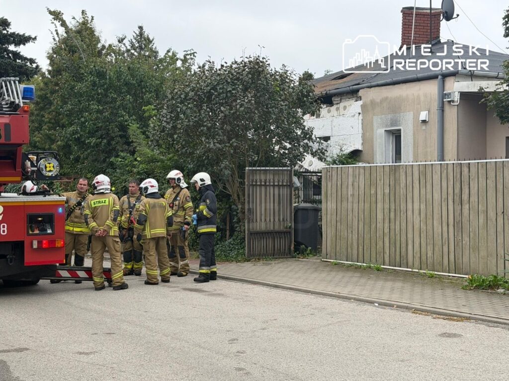 Grupa strażaków w mundurach omawia sytuację przed uszkodzonym domem, obok wozu strażackiego.