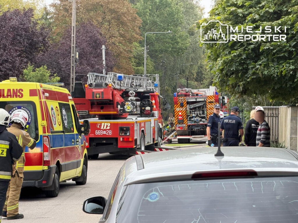 Strażacy w mundurach oraz pojazdy ratunkowe na ulicy podczas akcji gaśniczej po pożarze domu.