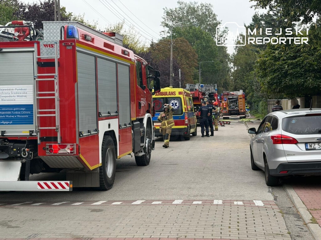 Wozidła strażackie i karetka na ulicy, a strażacy przygotowują się do akcji gaśniczej.