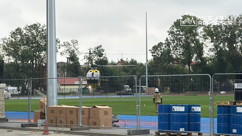 Helikopter ratunkowy ląduje na boisku sportowym, obok którego stoi strażak w pełnym umundurowaniu.