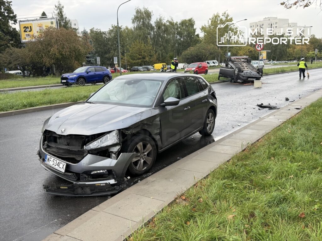 Zniszczony samochód osobowy stoi na poboczu, obok przewrócone auto leży na dachu, a obok pracują ratownicy.