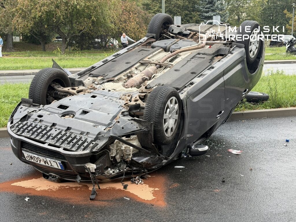 Samochód osobowy leży na dachu na jezdni, obok widoczne plamy płynów eksploatacyjnych i fragmenty szkła.
