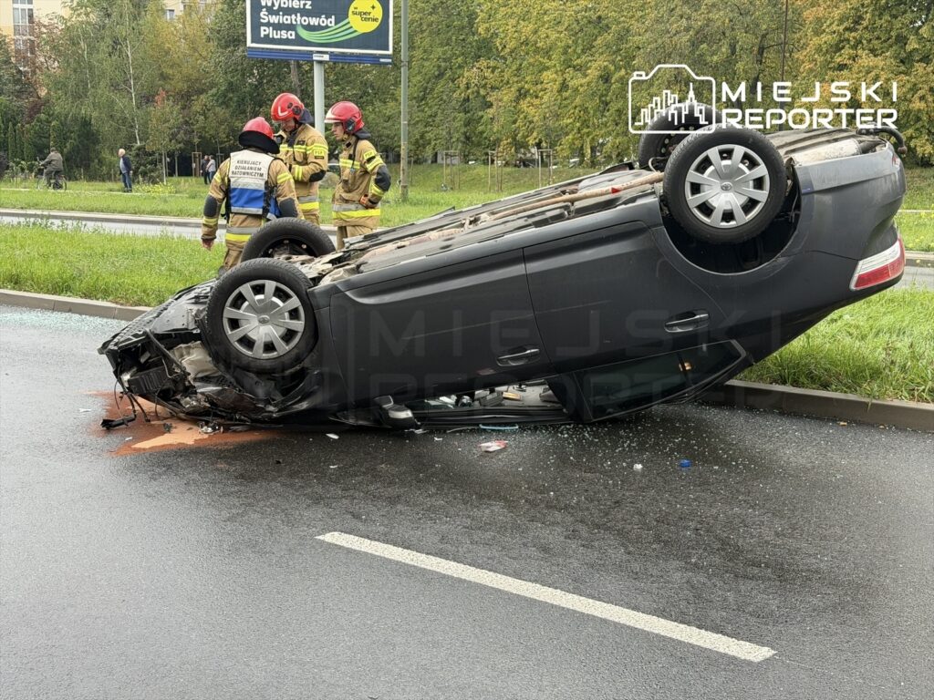 Czarny samochód osobowy leży na dachu na jezdni, obok trzech strażaków w hełmach, którzy oceniają sytuację.