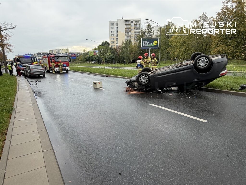 Na jezdni leży przewrócony samochód osobowy, obok niego pracują strażacy, a w tle widoczne są inne pojazdy i budynki.