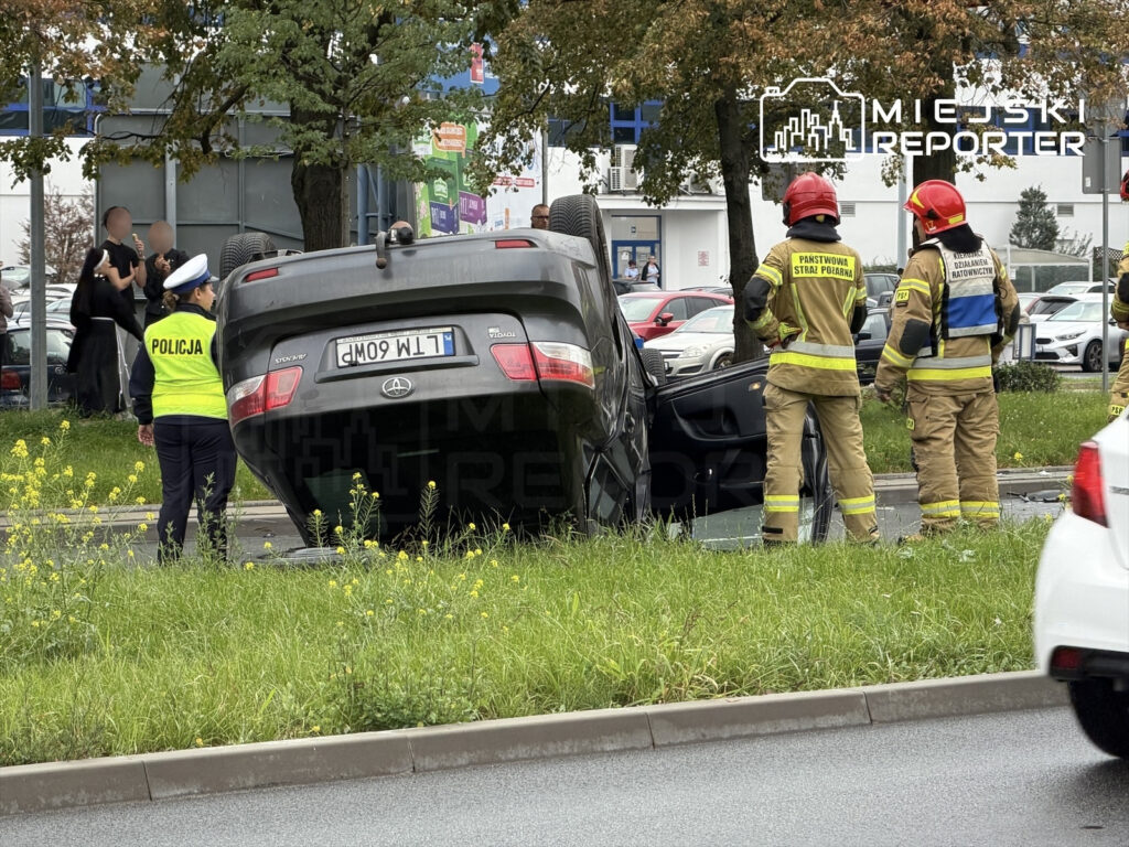 Na drodze leży przewrócone na dach auto, a obok stoją policjantka w kamizelce odblaskowej oraz strażacy w mundurach.