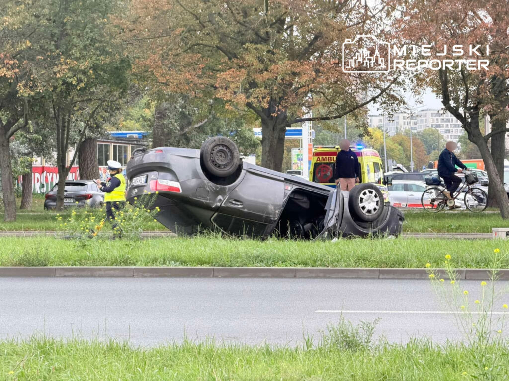 Czarny samochód osobowy leży na dachu na trawniku przy drodze, a policjant w żółtej kamizelce zabezpiecza miejsce zdarzenia.