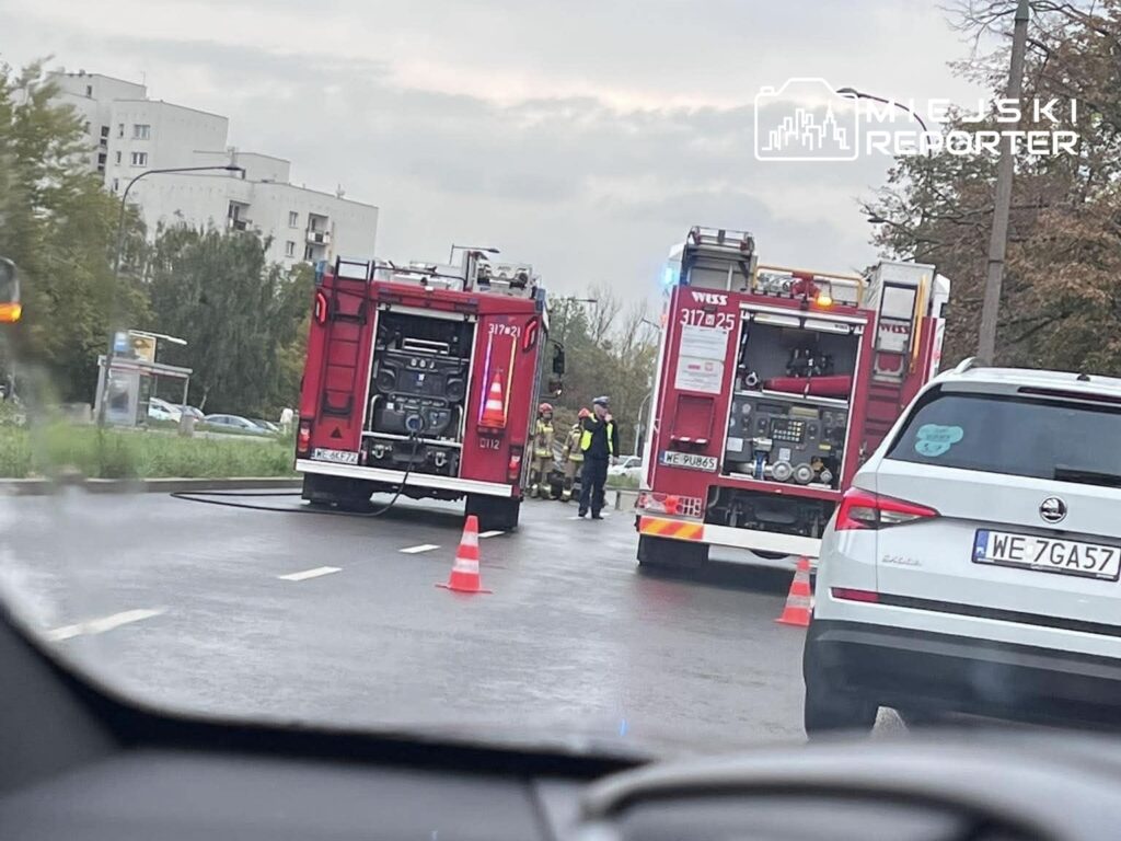 Dwa wozy strażackie z włączonymi sygnałami świetlnymi w trakcie akcji ratunkowej na ulicy, obok stojący strażacy.