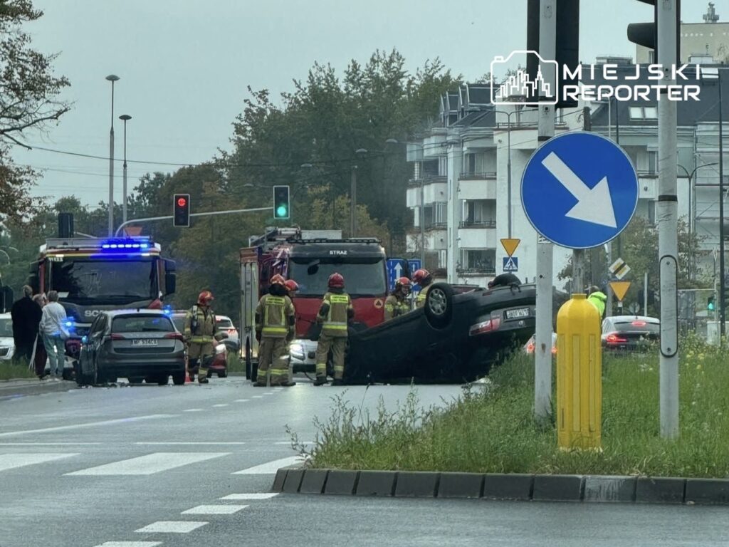 Na jezdni leży przewrócony samochód, obok niego interweniują strażacy w hełmach. W tle widać sygnalizację świetlną i inne pojazdy.