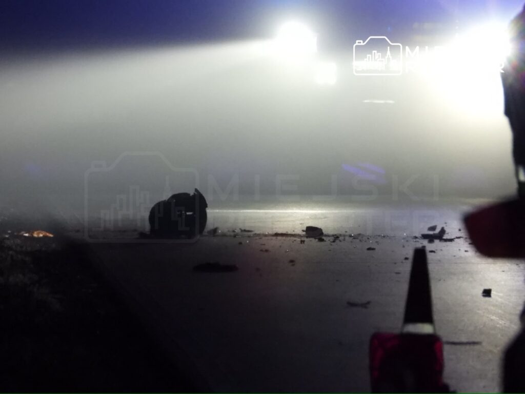 Na drodze leży przewrócona opona oraz fragmenty pojazdów, w tle widoczne światła policyjne w gęstej mgle.