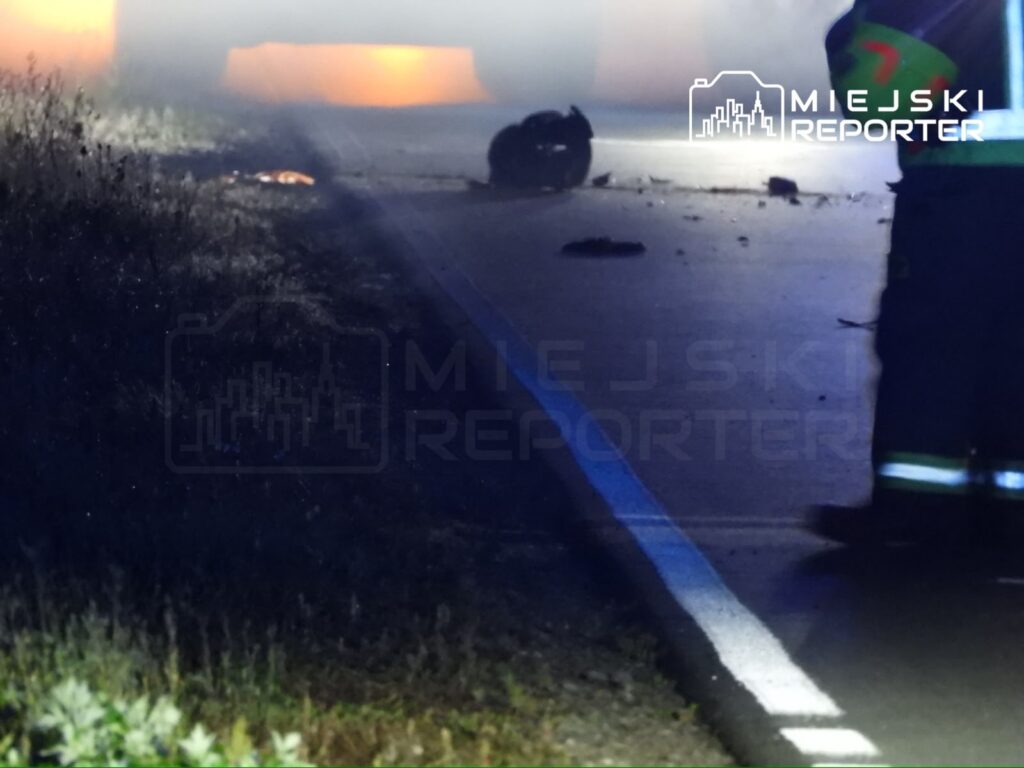 Na drodze, w mgle, leży wrak pojazdu, a w tle widać pojazd służb ratunkowych. Wokół rozrzucone fragmenty.