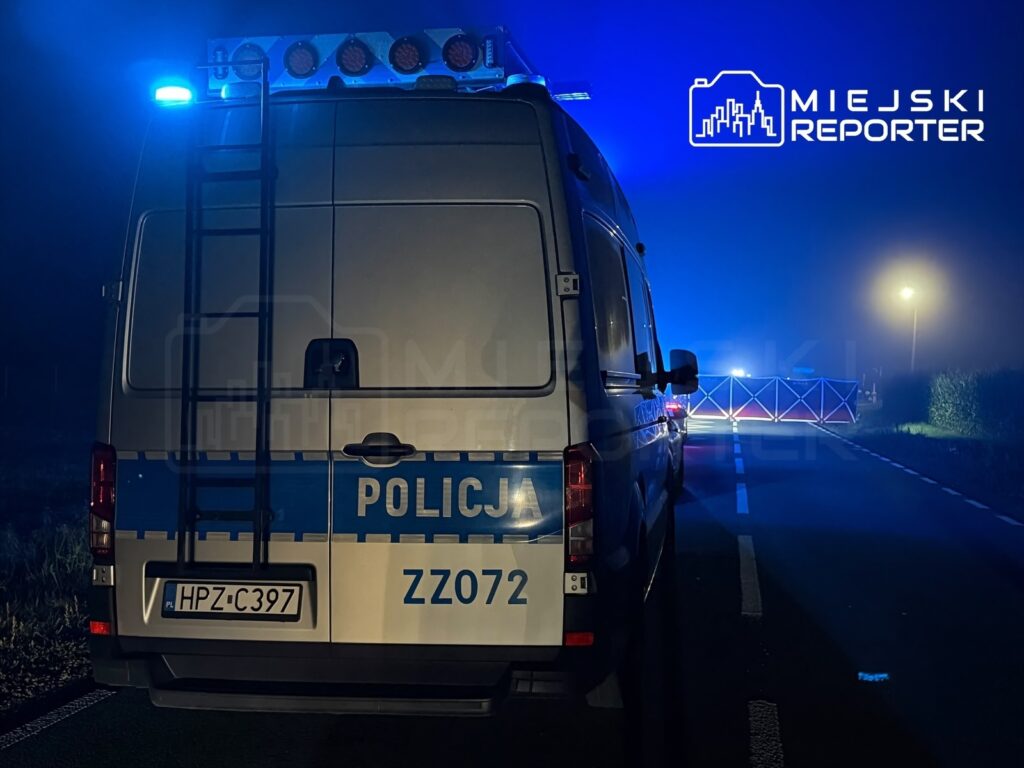 Policyjny radiowóz z włączonymi sygnałami świetlnymi zatrzymany na drodze, otoczony barierkami zabezpieczającymi w nocy.