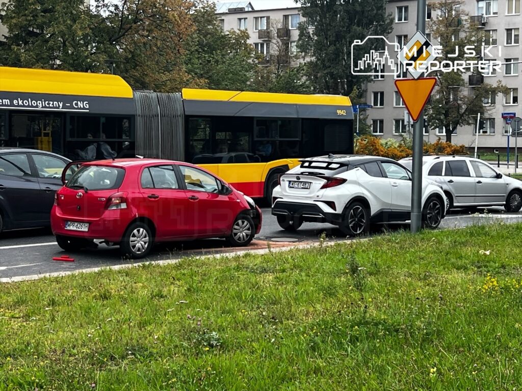 Czerwony samochód osobowy zderzony z białym SUV-em na ulicy obok przystanku autobusowego. W tle bloki mieszkalne.