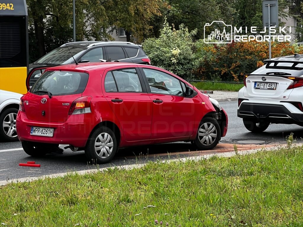 Czerwony samochód osobowy z uszkodzoną przednią częścią stoi na poboczu drogi, obok innych pojazdów.