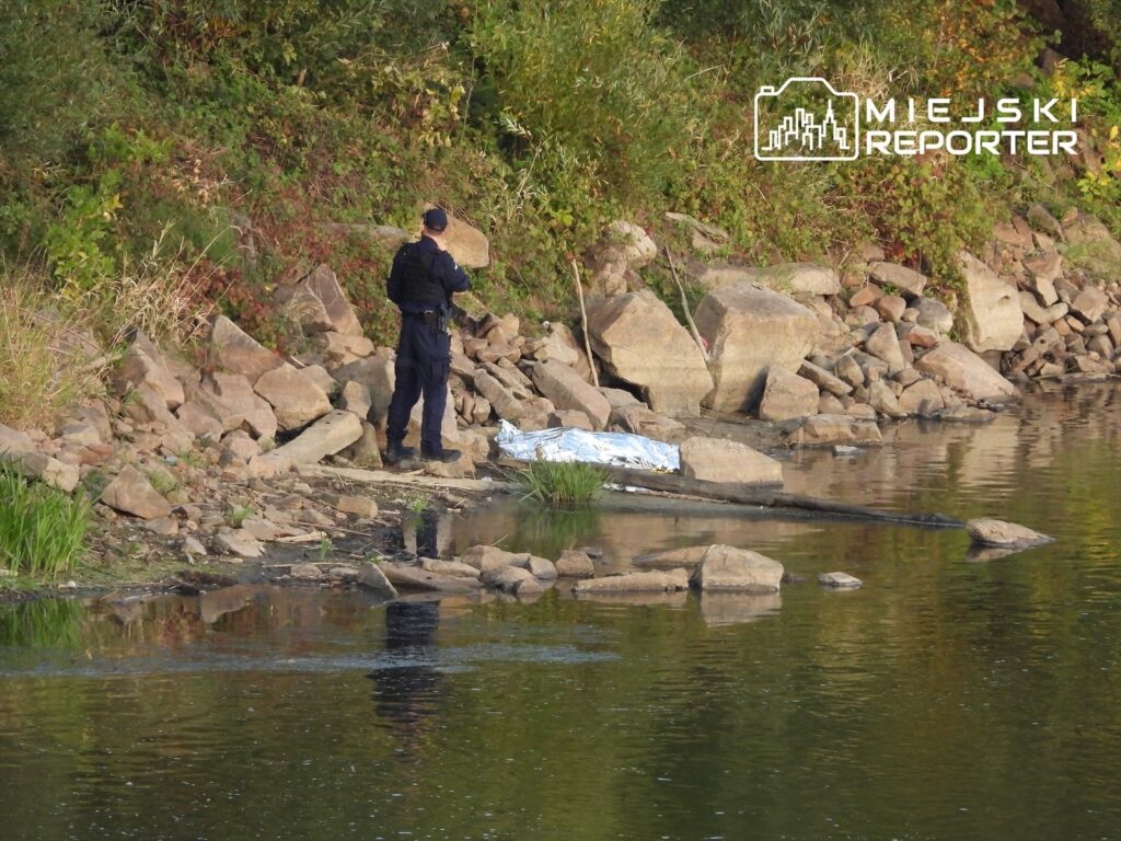 Funkcjonariusz policji bada miejsce zdarzenia na brzegu rzeki, obok leży przykryte ciało.